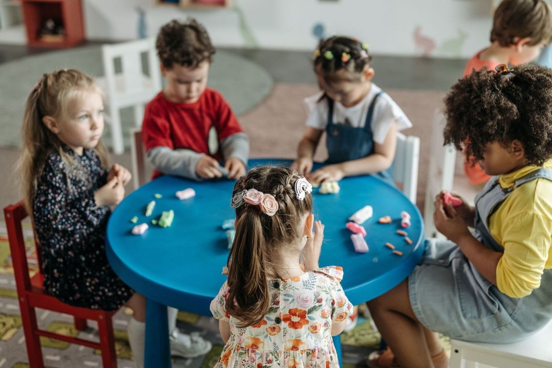 Children seated around a blue table, playing with colorful pieces in a classroom-like setting