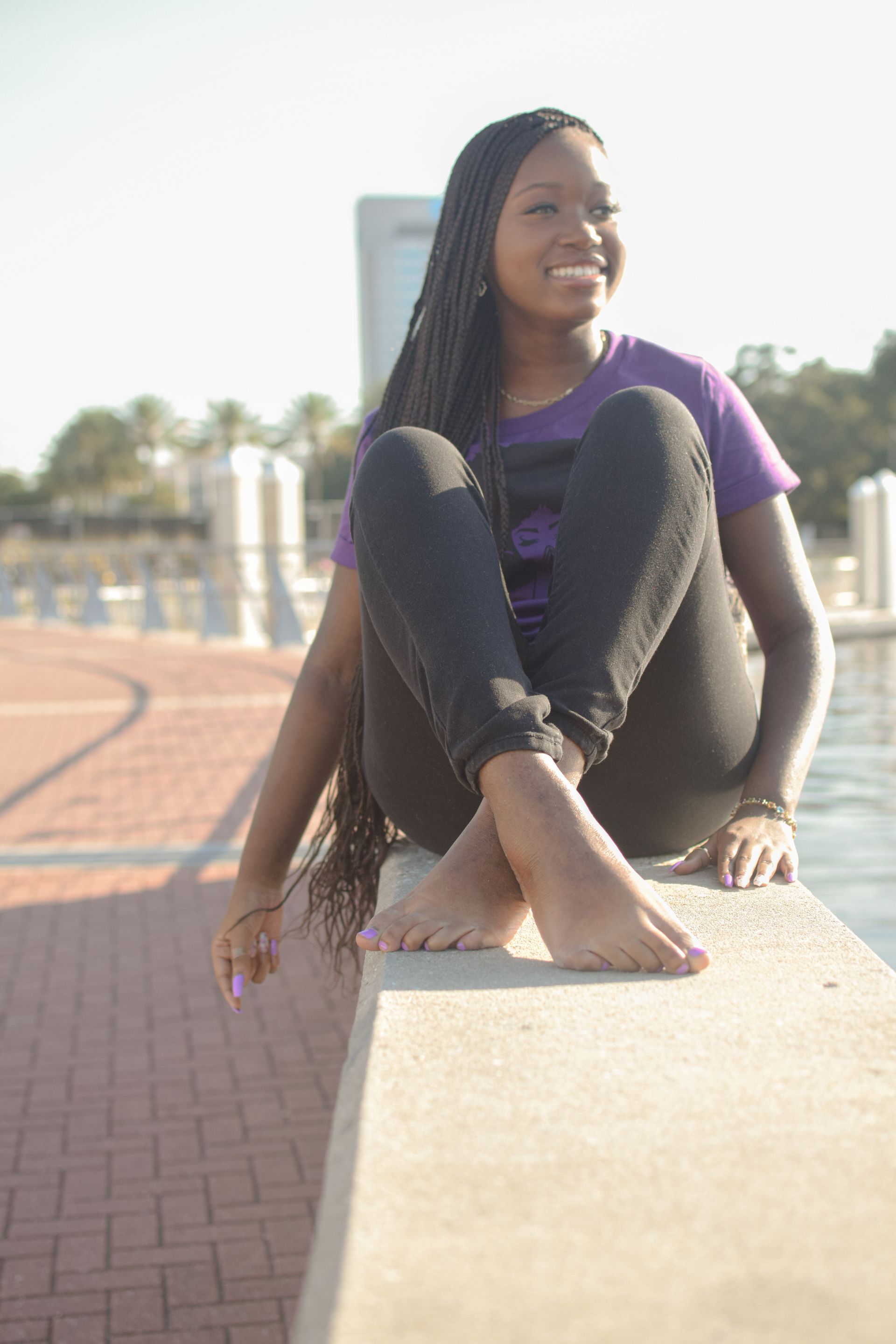 Young Black woman with long braids sits barefoot on a concrete ledge, smiling at something off-camera.