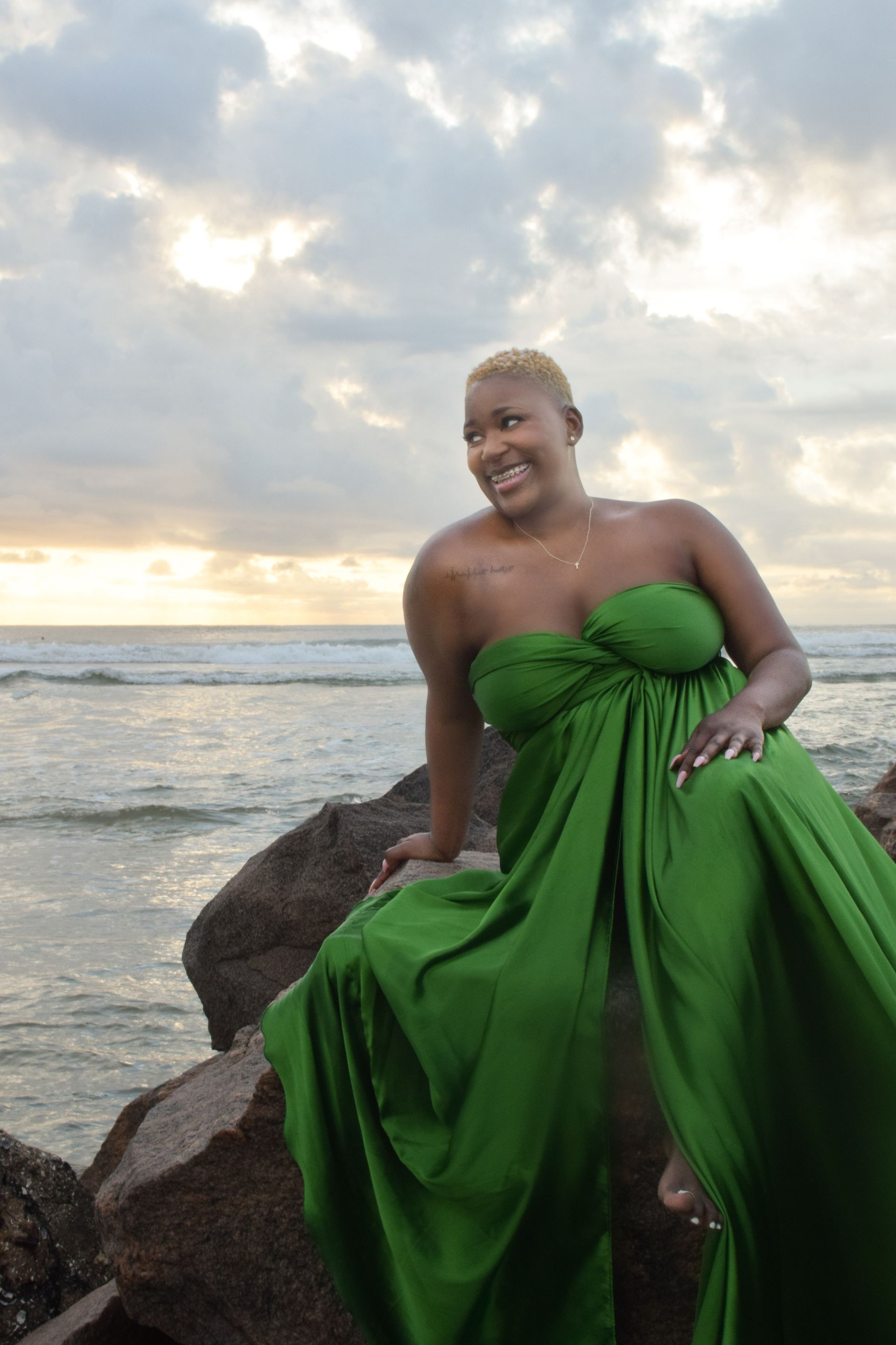 Woman in green dress on rocks by ocean, smiling at sunset.