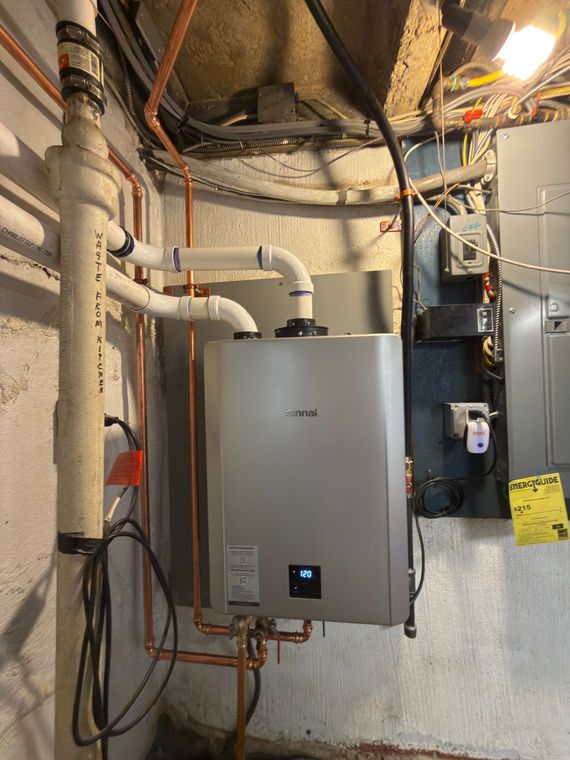 Tankless water heater with copper pipes, white PVC pipes, and electrical components against a concrete wall.