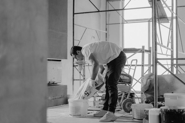 Person in white shirt and dark pants pouring from a bag into a bucket in a construction setting.
