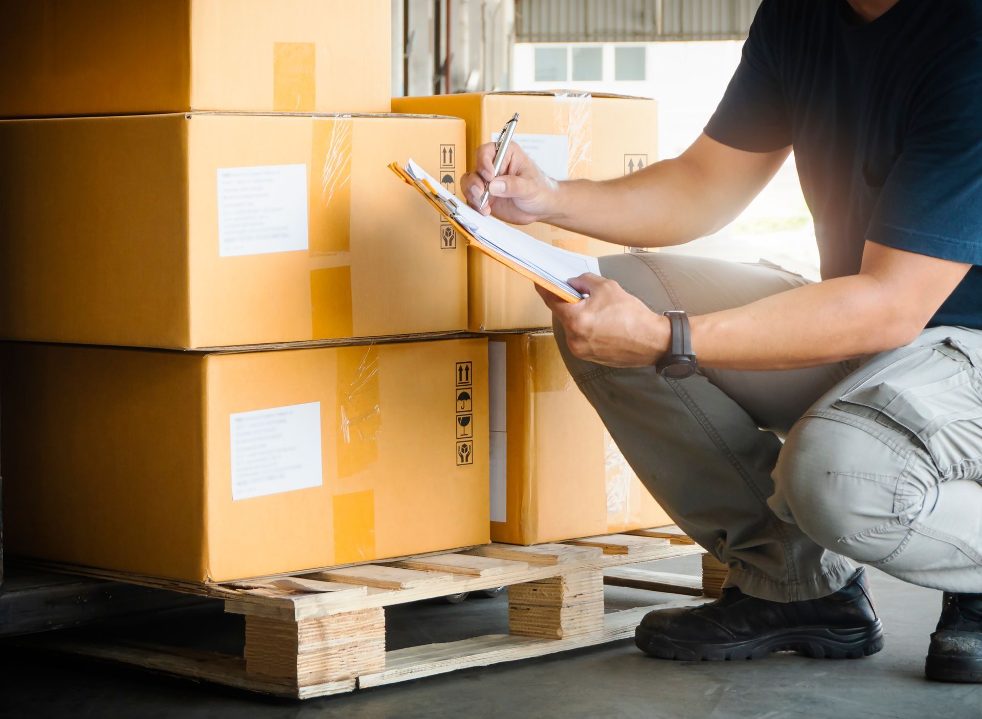 A man is kneeling down in front of a pallet of boxes and writing on a clipboard.