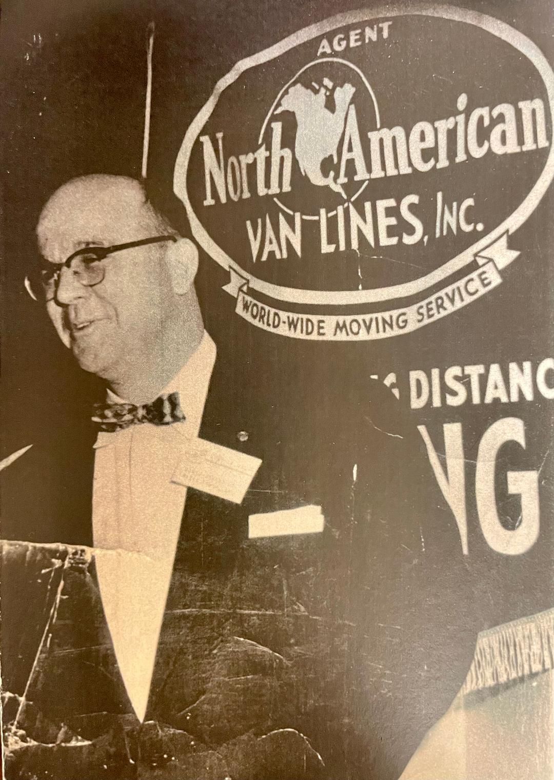 A man in a tuxedo stands in front of a sign for north america van lines inc.