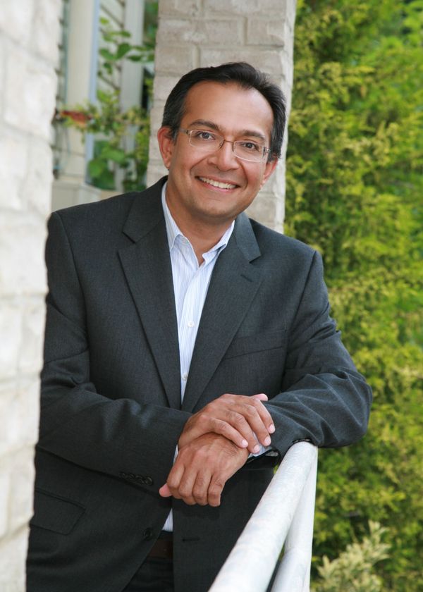 A man in a suit and glasses is leaning on a railing and smiling.