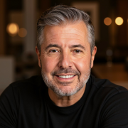Smiling man with graying hair in a black shirt, close-up portrait. Blurred background.