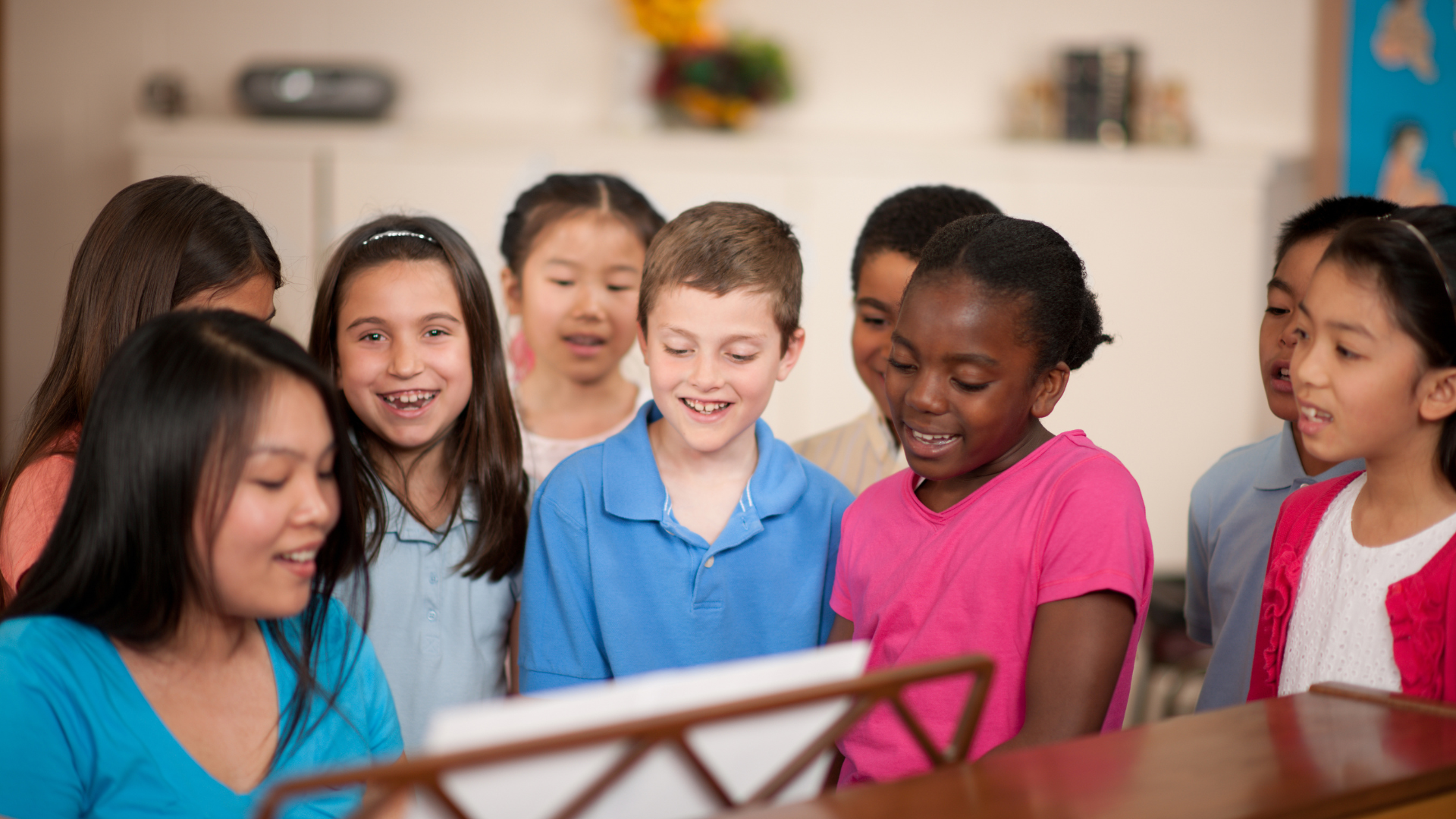 Children singing together around a piano, diverse group, happy expressions, classroom setting.