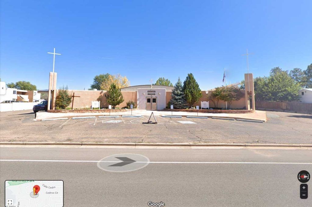 A one-story beige building with a cross and a flag outside, set against a blue sky.