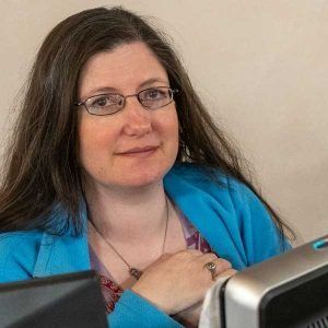 Woman with glasses, blue blazer, looking at camera, seated near a computer.