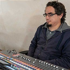 Man with glasses operating a soundboard, wearing a blue zip-up hoodie, in a studio setting.