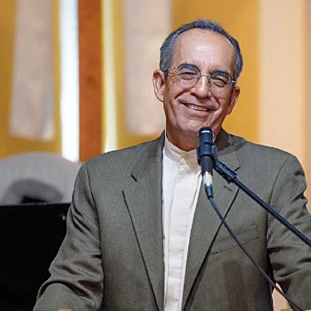 Smiling man in glasses speaks into microphone, wearing a suit jacket and light shirt.