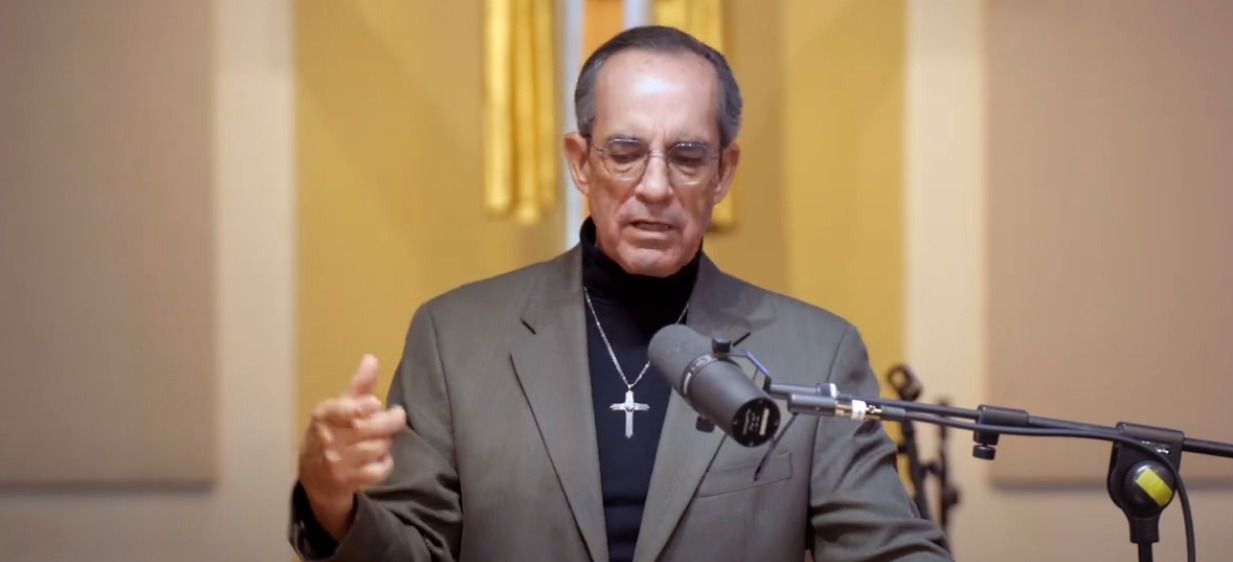 Man wearing glasses, a cross necklace, and a suit jacket, speaking into a microphone.