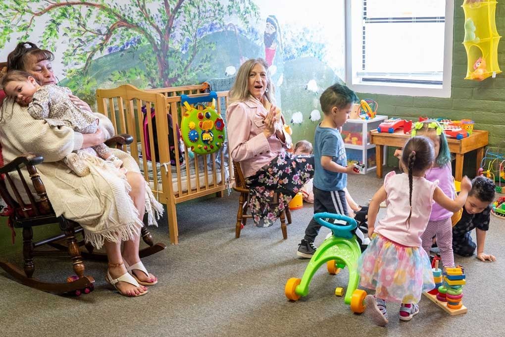 Childcare room: woman holding baby, woman seated, children playing with toys, crib, colorful decor.