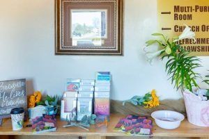 Wooden table with flowers, brochures, and coffee. Includes a sign indicating a church's multi-purpose room and office.