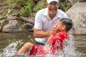 Man baptizing a young boy in a river. The boy is in red, splashing water. The man is in white, smiling.
