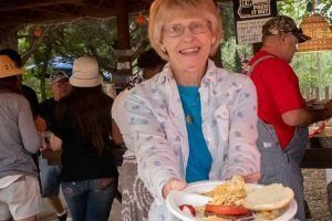 Woman smiling, holding plate of food at outdoor gathering. Other people visible in background.
