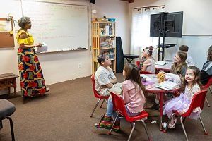 Teacher in colorful dress leading a lesson for children seated at a small round table; whiteboard visible.