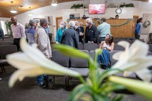 People gather in a church. Foreground has white lilies, and the room has gray chairs.
