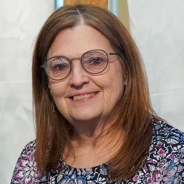 Woman with glasses, smiling, wearing a patterned top. Light background.
