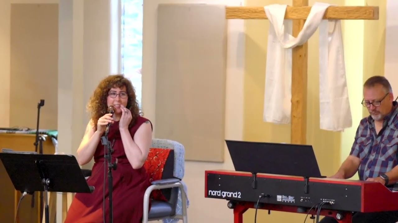 Woman singing, red dress, holding microphone, by keyboard player. Church setting, cross on wall.