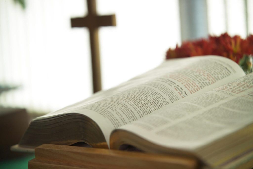 Open Bible on a stand in front of a wooden cross in a church setting.