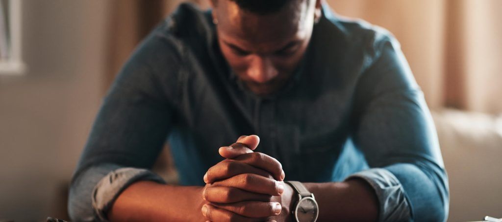 Man with clasped hands, looking down. Wearing a denim shirt and a watch.