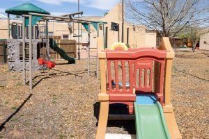 Playground equipment in a gravel yard; a red and tan playhouse with a green slide, a gray climbing structure, and swings.