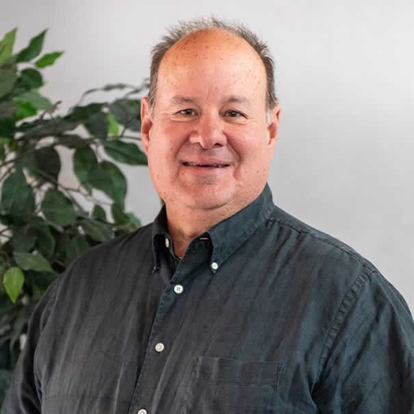 Middle-aged man in dark shirt smiles at the camera, with a plant in the background.