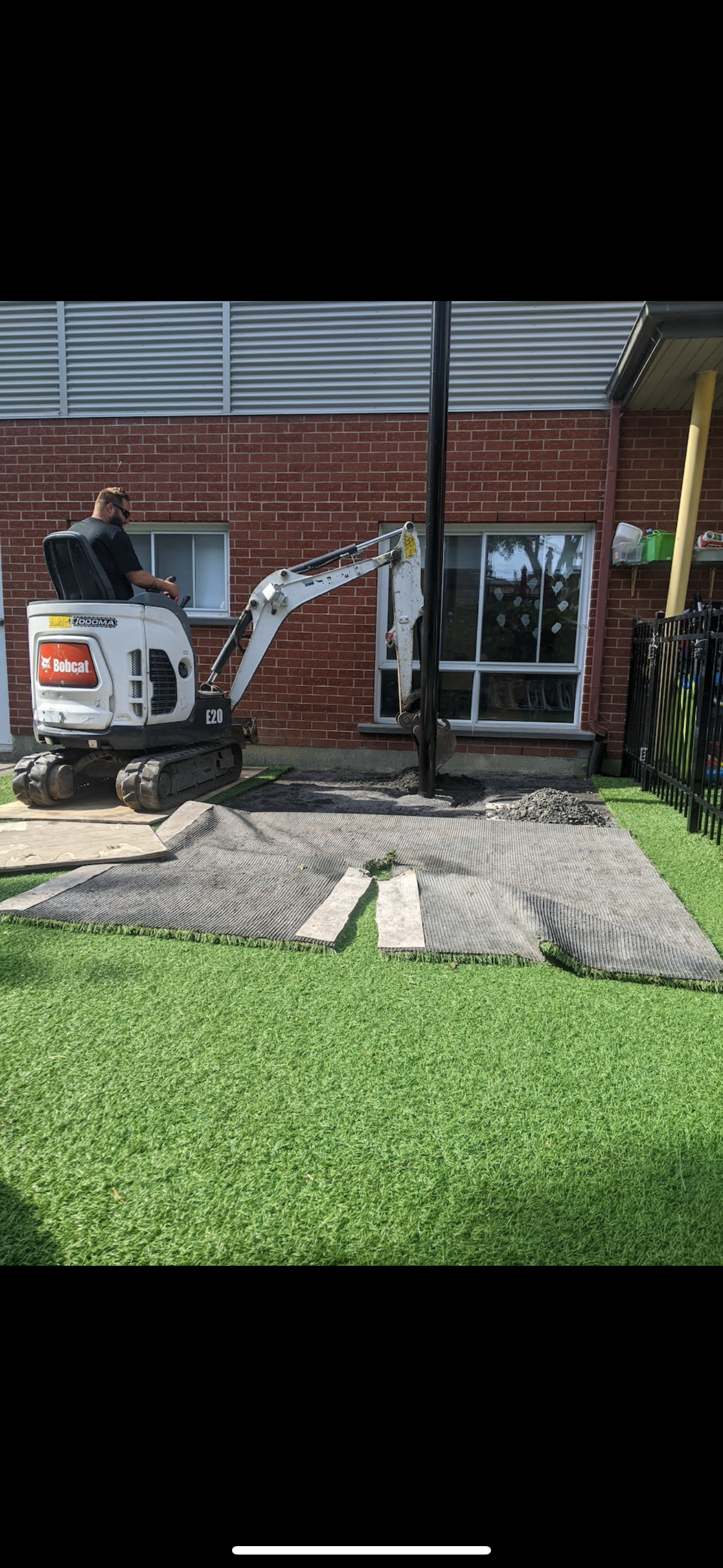 Un homme conduit une petite excavatrice devant un bâtiment en briques.