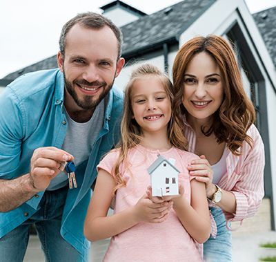 Family smiling, holding house model and keys in front of a new home.