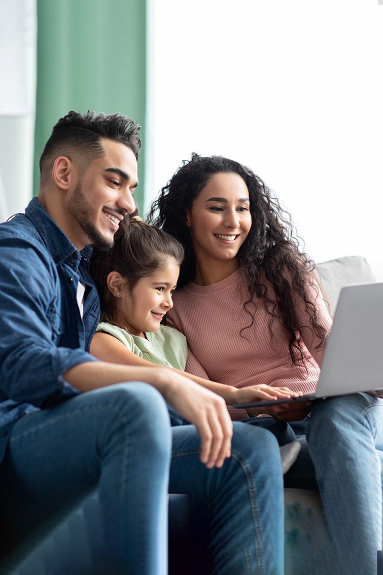 Family of three smiling, looking at a laptop together on a couch.