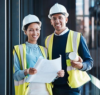 Two construction workers in hard hats and vests smiling, holding blueprints and a cup.