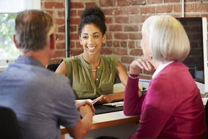 Woman smiling, talking to a couple at a desk in an office with a brick wall.