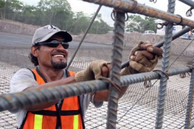 Construction worker smiling, securing rebar structure with gloves, wearing vest and cap.