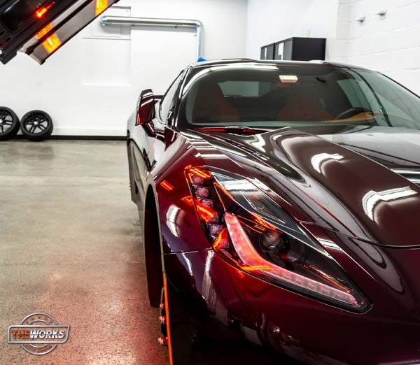 Maroon sports car in a garage with bright lights; focused on the car's front with lights on.