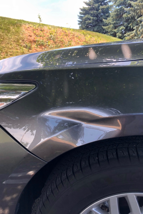 Gray car with a large dent in the front fender. Tire visible below.