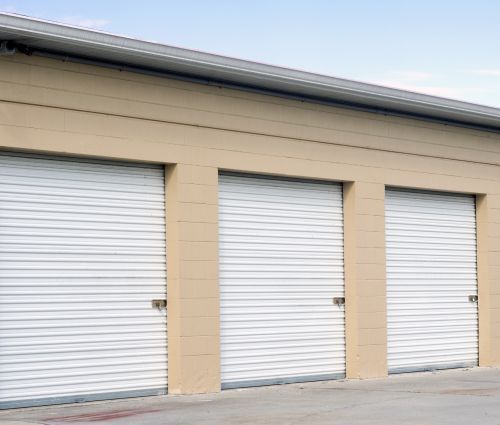 Row of white, closed storage unit doors with beige building and blue sky.