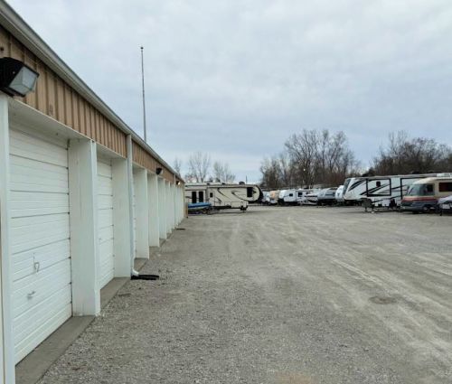 Storage units with white doors, gravel lot, and RVs parked in the background under an overcast sky.
