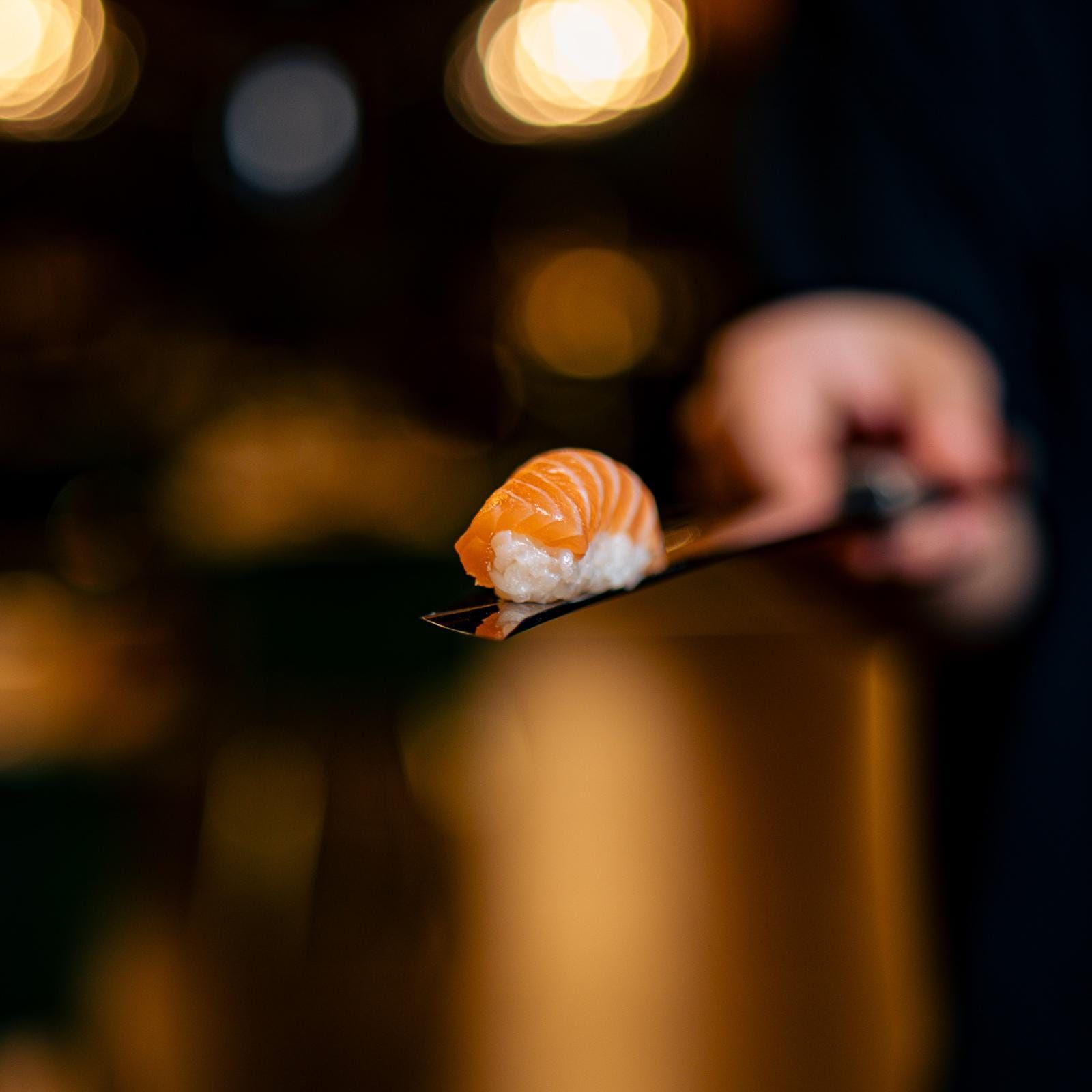 Sushi nigiri di salmone su un coltello, tenuto in mano. Luci bokeh sullo sfondo.