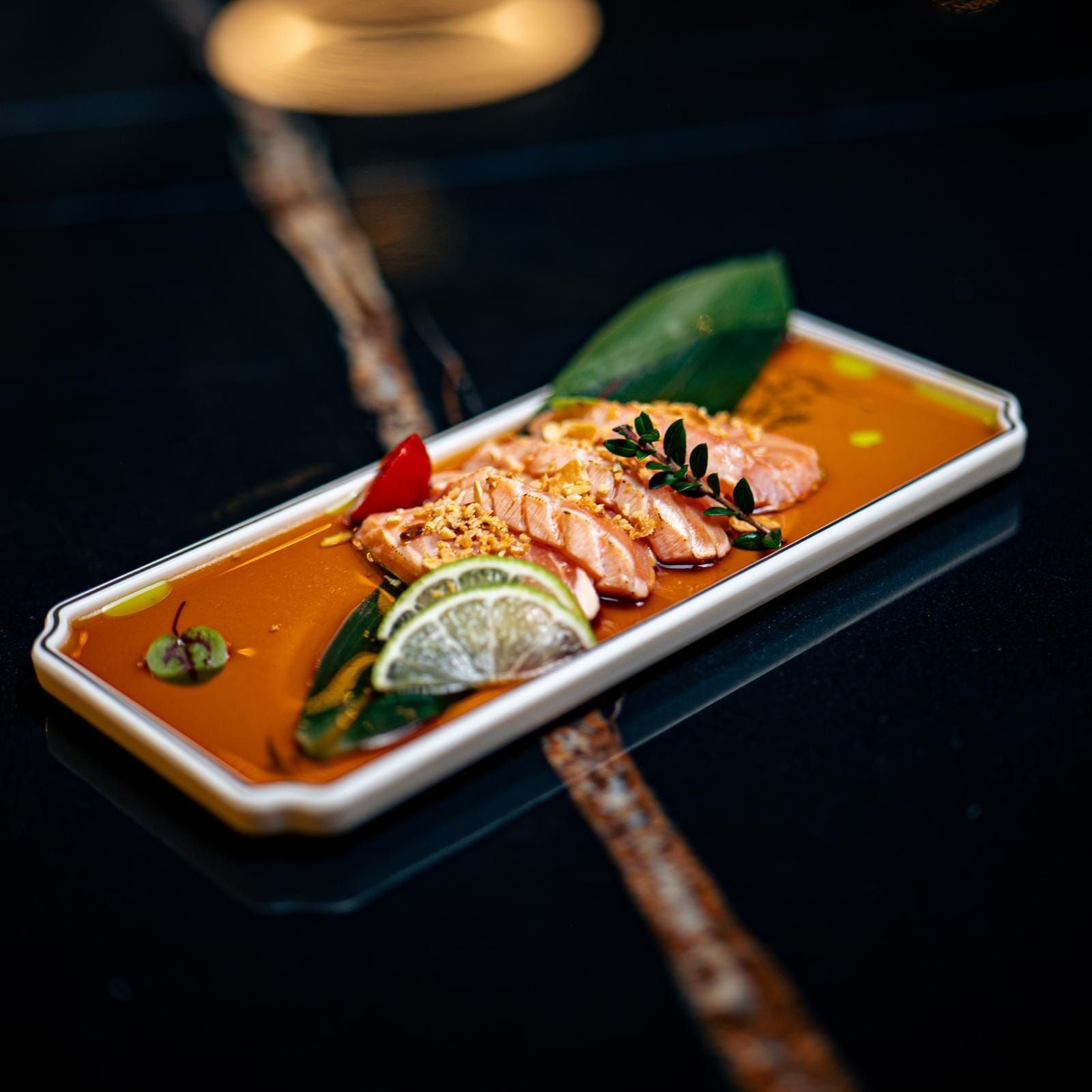Sashimi di salmone su piatto rettangolare con salsa, lime, verdure e guarnizione su superficie nera.