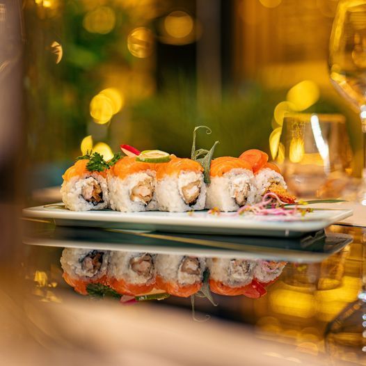 Rotoli di sushi su un piatto bianco, riflessi sotto su una superficie lucida, sfondo bokeh caldo.