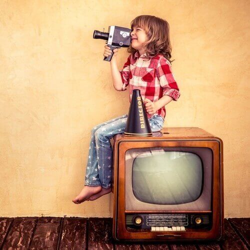 A little girl is sitting on a television holding a camera and a megaphone.