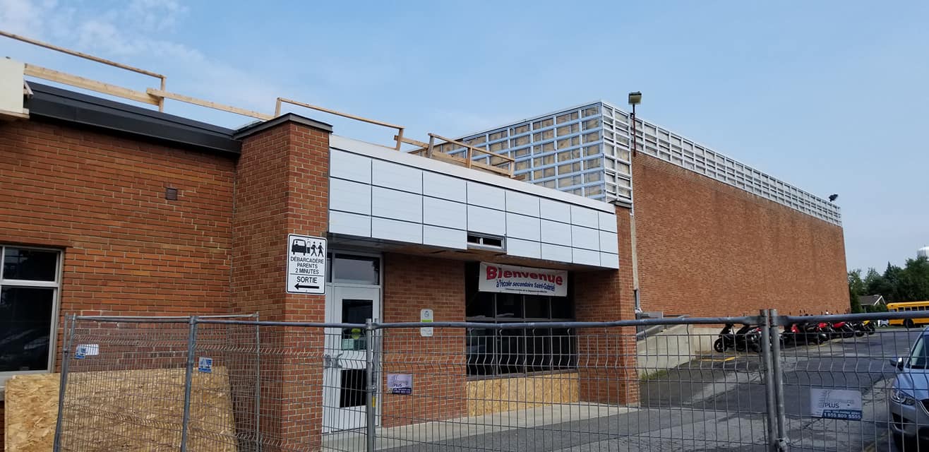 Extérieur d'un bâtiment en briques en construction, avec revêtement métallique et enseigne. Zone clôturée.