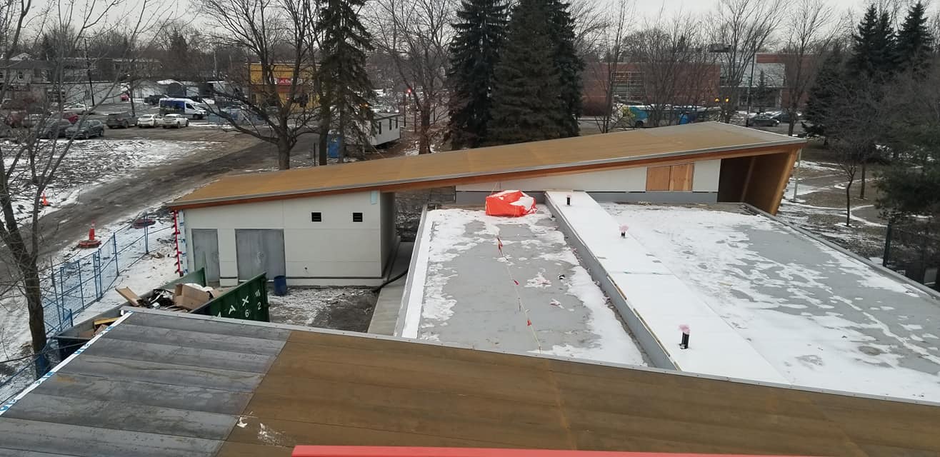 Des bâtiments aux toits bruns et aux toits plats recouverts de neige, des arbres, une route et un ciel d'hiver nuageux.