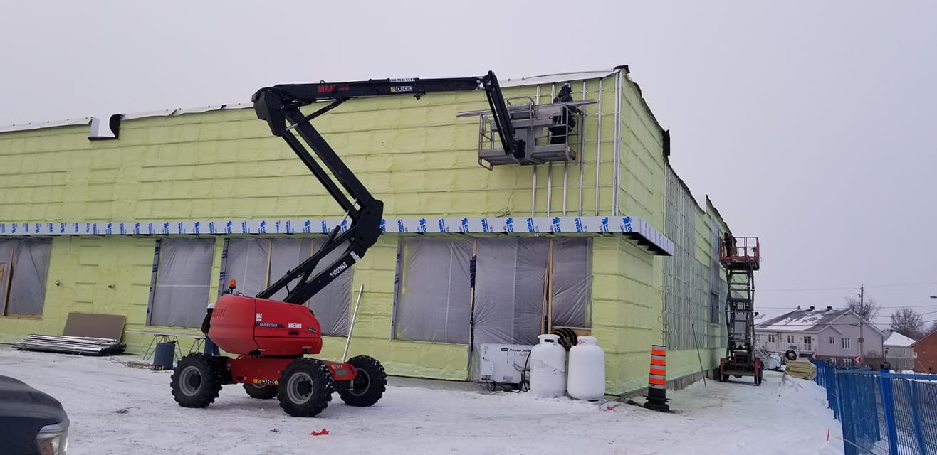 Une nacelle élévatrice rouge près d'un bâtiment avec des ouvriers dans la nacelle. Sol enneigé.