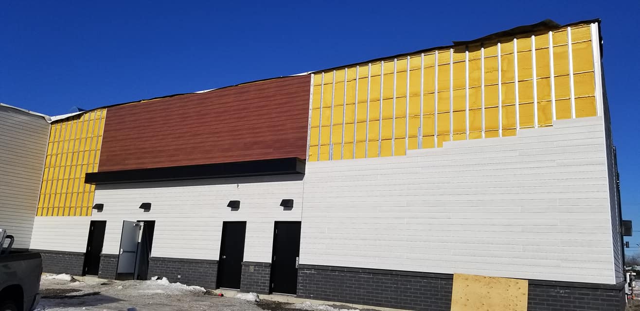 Extérieur d'un bâtiment en construction, montrant différents matériaux de revêtement : toit rouge, revêtement blanc, isolation apparente et base en brique.