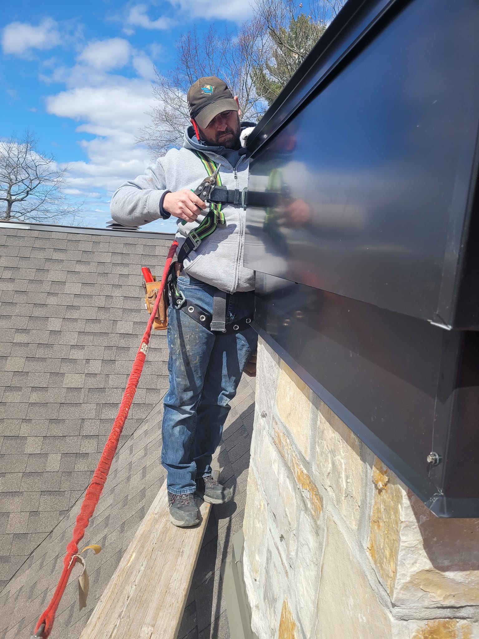Ouvrier du bâtiment sur un toit portant un harnais de sécurité, travaillant sur des solins en métal foncé.