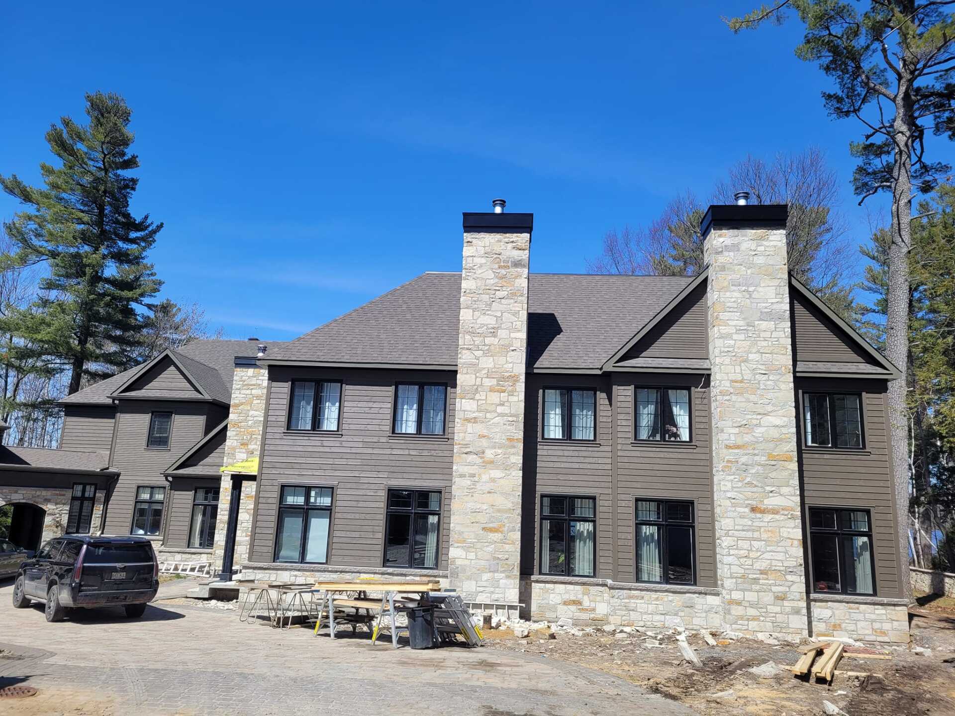 Grande maison à deux étages avec cheminées en pierre et revêtement brun sous un ciel bleu clair.