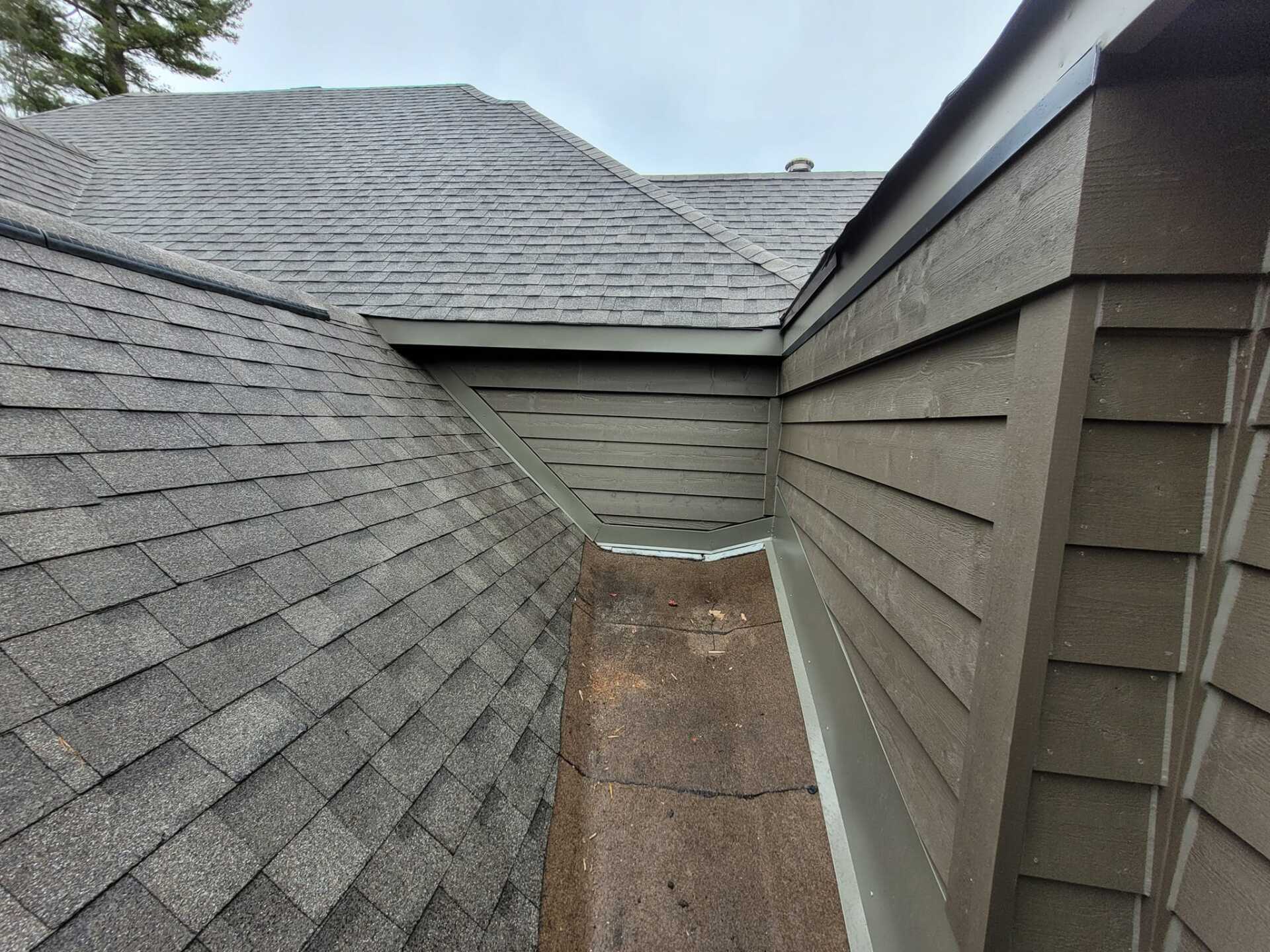 Toiture en bardeaux d'asphalte gris rencontrant un mur de revêtement brun avec une gouttière, ciel couvert.