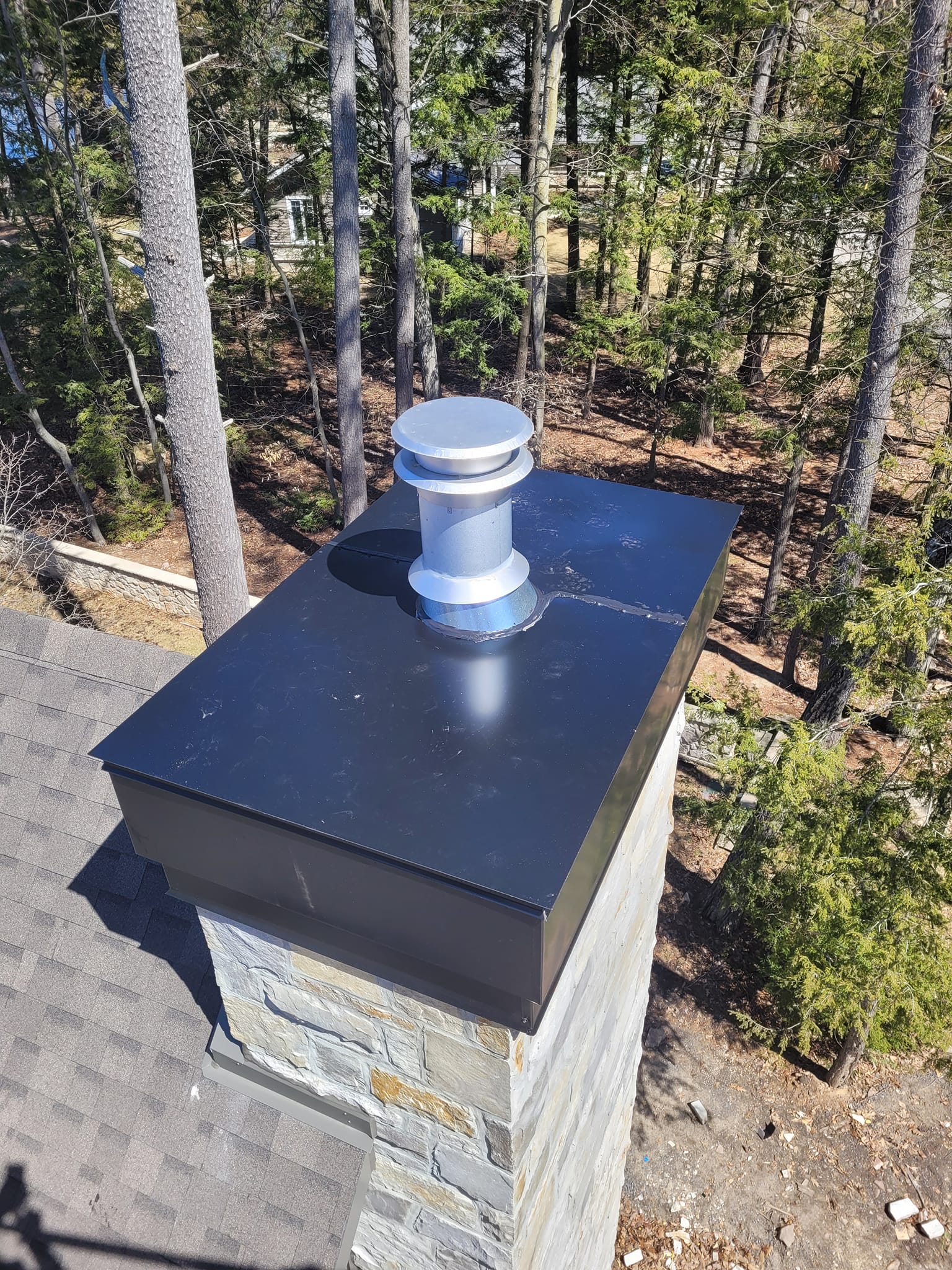 Cheminée avec un chapeau en métal noir et un évent sur un toit en bardeaux, arbres en arrière-plan.