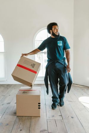 A person in a dark green t-shirt stands on a wood floor, placing a cardboard box on top of another in a bright room.